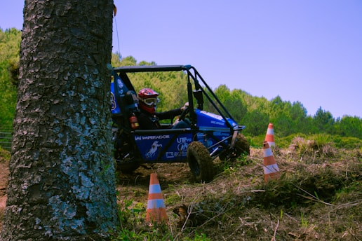A blue off-road buggy is maneuvering through a natural, uneven landscape. The vehicle is partially obscured by a large tree on the left side of the image. The driver, wearing a helmet and protective gear, is focused on steering the buggy. Two orange traffic cones are visible on the ground, indicating a marked path. Dense green foliage and trees fill the background, suggesting a forest setting, under a clear blue sky.