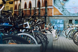 A row of parked bicycles lines a cobblestone street near a brick building with arched windows. The foreground features close-up details of bicycle frames, tires, and handlebars, while the background shows an urban setting with billboards and other distant bicycles.