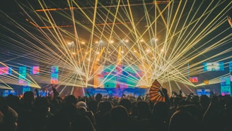 A vibrant concert scene with a large crowd enjoying the performance. Multiple lasers in shades of green, blue, and yellow create a dynamic light show above the audience. The stage is illuminated with colorful spotlights and displays on screens.