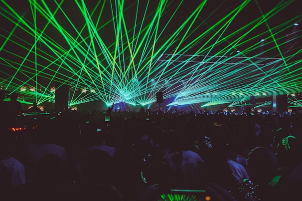 A vibrant nightclub or concert scene with numerous green laser beams radiating over a large crowd. The lasers create a dynamic and energetic atmosphere, reflecting off various surfaces and illuminating the space. The crowd appears engaged and animated, possibly taking photos or recording videos with their phones.