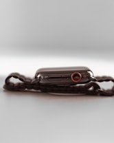 A sleek, modern smartwatch with a dark, polished metal casing and a black chain link strap is shown. The crown of the watch features a prominent red accent. The watch is placed on a reflective, light-colored surface, creating a minimalistic and sophisticated appearance.