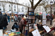 An outdoor art market with painters displaying their work. Various pieces of art are set up on easels, and there are several people dressed in winter clothing browsing and interacting. Trees without leaves are in the background, and buildings with classic architecture line the street.