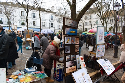 An outdoor art market with painters displaying their work. Various pieces of art are set up on easels, and there are several people dressed in winter clothing browsing and interacting. Trees without leaves are in the background, and buildings with classic architecture line the street.