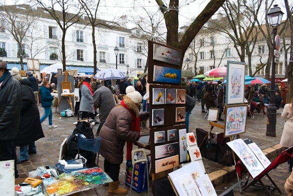 An outdoor art market with painters displaying their work. Various pieces of art are set up on easels, and there are several people dressed in winter clothing browsing and interacting. Trees without leaves are in the background, and buildings with classic architecture line the street.
