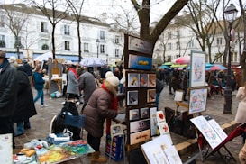 An outdoor art market with painters displaying their work. Various pieces of art are set up on easels, and there are several people dressed in winter clothing browsing and interacting. Trees without leaves are in the background, and buildings with classic architecture line the street.