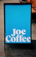 A blue signboard with bold white text reading 'Joe Coffee' placed on a sidewalk, with a wooden bench visible in the background.