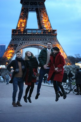 A group of four people is joyfully jumping in front of the illuminated Eiffel Tower in the evening. They are wearing winter clothes, with two women and two men appearing happy and cheerful. The background shows a clear view of the Eiffel Tower, lit up against a dusk sky with a few visible clouds.