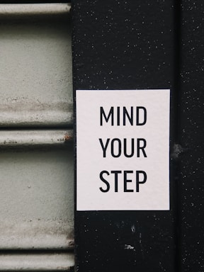 A white sign with black text reading 'MIND YOUR STEP' is affixed to a black surface. To the left of the sign, a textured vertical surface in a light grayish-green color is visible.