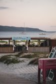 A beachside setup with a wooden bar area named 'Porto Pino' in the foreground. Green and yellow beach umbrellas are arranged behind the bar area. There are boats and yachts anchored in the calm sea. In the background, there are rolling hills under a lightly clouded sky. To the right, there is a stack of chairs.