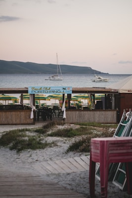 A beachside setup with a wooden bar area named 'Porto Pino' in the foreground. Green and yellow beach umbrellas are arranged behind the bar area. There are boats and yachts anchored in the calm sea. In the background, there are rolling hills under a lightly clouded sky. To the right, there is a stack of chairs.