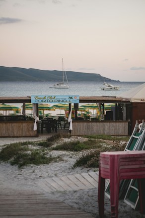 A beachside setup with a wooden bar area named 'Porto Pino' in the foreground. Green and yellow beach umbrellas are arranged behind the bar area. There are boats and yachts anchored in the calm sea. In the background, there are rolling hills under a lightly clouded sky. To the right, there is a stack of chairs.