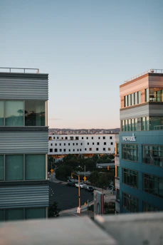 Two modern multi-story office buildings flank a street, with the sun casting warm light on their structures. The sky is clear with soft pastel hues, and a white building is visible in the background. Cars are parked along the street, and a few trees add greenery to the urban landscape.