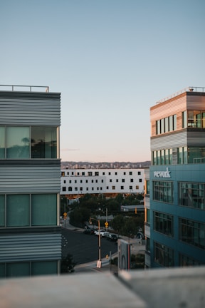 Two modern multi-story office buildings flank a street, with the sun casting warm light on their structures. The sky is clear with soft pastel hues, and a white building is visible in the background. Cars are parked along the street, and a few trees add greenery to the urban landscape.