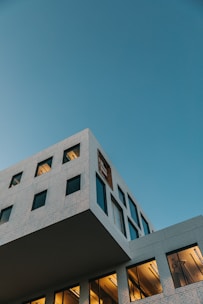 A modern, angular building with a minimalist design, featuring large windows and light brickwork set against a clear blue sky. Warm light emanates from the lower windows, creating a contrast with the cooler tones of the sky and building exterior.