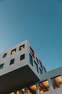 A modern, angular building with a minimalist design, featuring large windows and light brickwork set against a clear blue sky. Warm light emanates from the lower windows, creating a contrast with the cooler tones of the sky and building exterior.