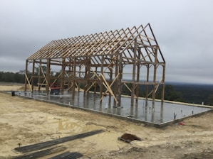 A wooden framework of a large structure stands on a newly poured concrete foundation. The surrounding area appears to be a construction site with sand, dirt, and metal bars scattered around. The sky is overcast, indicating a cloudy day, and there are distant trees visible in the background.