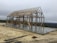 A wooden framework of a large structure stands on a newly poured concrete foundation. The surrounding area appears to be a construction site with sand, dirt, and metal bars scattered around. The sky is overcast, indicating a cloudy day, and there are distant trees visible in the background.