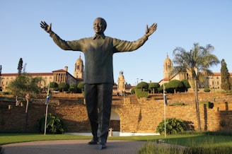 A large statue of a figure with open arms stands prominently in front of an impressive government building surrounded by well-maintained gardens. The structure features ornate architecture with columns and towers, and the landscape includes various trees and a stone wall.