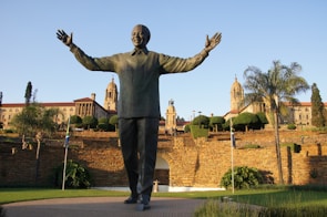 A large statue of a figure with open arms stands prominently in front of an impressive government building surrounded by well-maintained gardens. The structure features ornate architecture with columns and towers, and the landscape includes various trees and a stone wall.
