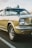 A classic beige Ford Mustang with chrome detailing and distinctively styled wheels. The image captures the car from a low angle, highlighting its retro features and iconic shape. The background shows a blurred outdoor setting, and the lighting suggests a late afternoon ambiance.