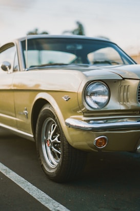 A classic beige Ford Mustang with chrome detailing and distinctively styled wheels. The image captures the car from a low angle, highlighting its retro features and iconic shape. The background shows a blurred outdoor setting, and the lighting suggests a late afternoon ambiance.