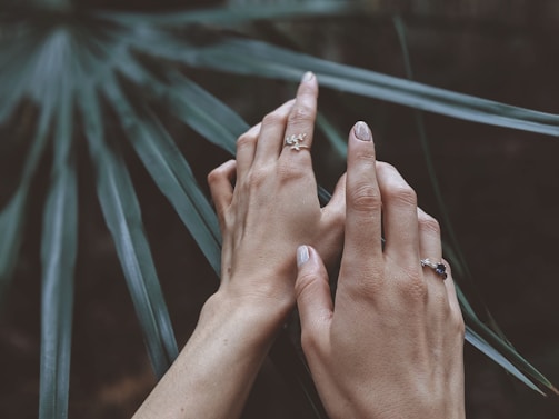 Two hands with rings are gently placed on large green palm leaves. The hands are well-manicured and adorned with delicate jewelry.