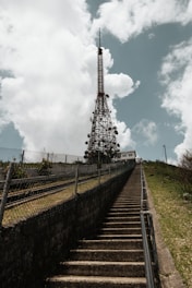 A tall metal communication tower stands against a background of fluffy clouds and a blue sky. A set of concrete stairs leads up to the tower, flanked by metal railings and a wire fence, with greenery visible on the sides.