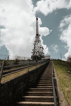 A tall metal communication tower stands against a background of fluffy clouds and a blue sky. A set of concrete stairs leads up to the tower, flanked by metal railings and a wire fence, with greenery visible on the sides.