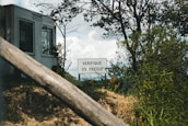 A white building stands in the background, partially obscured by trees and bushes. In front of it, there is a sign with the text 'VERIFIQUE OS FREIOS' surrounded by green foliage. The scene appears outdoors with a focus on nature and a slightly overcast sky.