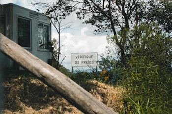 A white building stands in the background, partially obscured by trees and bushes. In front of it, there is a sign with the text 'VERIFIQUE OS FREIOS' surrounded by green foliage. The scene appears outdoors with a focus on nature and a slightly overcast sky.