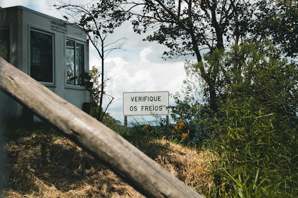 A white building stands in the background, partially obscured by trees and bushes. In front of it, there is a sign with the text 'VERIFIQUE OS FREIOS' surrounded by green foliage. The scene appears outdoors with a focus on nature and a slightly overcast sky.