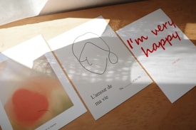 Three pieces of paper are displayed on a wooden surface. The left paper features an abstract design with peach and beige tones. The middle paper has a minimalist line drawing of a face with French text 'L'amour de ma vie' and a missing word fill-in. The right paper displays bold, red handwritten text stating 'I'm very happy'. Sunlight creates soft shadows on the papers.