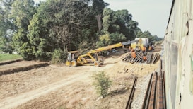 A construction scene by a railroad track with workers in orange uniforms operating machinery. A yellow crane is lifting steel beams beside a pair of tracks extending into the distance. Lush green trees line the side of the tracks, and a partially visible train moves on the right.