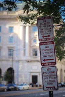 Multiple street signs indicate parking restrictions next to a busy urban street with a historic building in the background. The signs detail specific no-parking times during weekdays and restrictions for sightseeing buses.