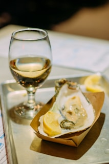 A single oyster is served on a half shell with a lemon wedge in a small paper tray, placed on a metal tray. Beside it, a glass of white wine is partially filled. The background is softly blurred, emphasizing the oyster and wine as the main focus.