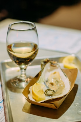 A single oyster is served on a half shell with a lemon wedge in a small paper tray, placed on a metal tray. Beside it, a glass of white wine is partially filled. The background is softly blurred, emphasizing the oyster and wine as the main focus.