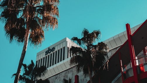 A modern building with a distinctive sbe logo on the upper section is surrounded by tall palm trees. The architectural design features sleek lines and a combination of white and gray tones with red accents. A clear blue sky provides a contrasting background.