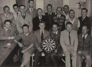 A group of fifteen men are gathered in a small room, with some standing and others seated around a table. They are dressed in various styles, including sweaters, jackets, and ties, reflecting mid-20th-century fashion. One man is holding a dartboard, indicating a possible club or team gathering.