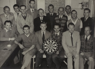 A group of fifteen men are gathered in a small room, with some standing and others seated around a table. They are dressed in various styles, including sweaters, jackets, and ties, reflecting mid-20th-century fashion. One man is holding a dartboard, indicating a possible club or team gathering.