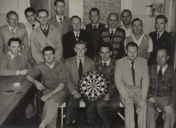 A group of fifteen men are gathered in a small room, with some standing and others seated around a table. They are dressed in various styles, including sweaters, jackets, and ties, reflecting mid-20th-century fashion. One man is holding a dartboard, indicating a possible club or team gathering.