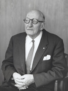 An elderly man with glasses is seated, wearing a suit and tie. He has a thoughtful expression and his hands are resting in his lap.