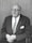 An elderly man with glasses is seated, wearing a suit and tie. He has a thoughtful expression and his hands are resting in his lap.
