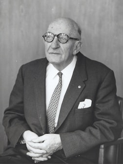 An elderly man with glasses is seated, wearing a suit and tie. He has a thoughtful expression and his hands are resting in his lap.