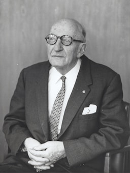 An elderly man with glasses is seated, wearing a suit and tie. He has a thoughtful expression and his hands are resting in his lap.