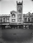 A historic street scene features an array of early 20th-century buildings with ornate architecture. Prominent signage includes 'Kodak House' on a tall building at the center and 'Taylor & Sharp' on the adjacent establishment. Other visible signs indicate businesses such as a dentist and health food shop. Overhead, power lines intersect the view, and a few pedestrians are present on the sidewalk. There is a bicycle parked near one of the shopfronts.