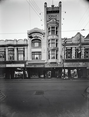 A historic street scene features an array of early 20th-century buildings with ornate architecture. Prominent signage includes 'Kodak House' on a tall building at the center and 'Taylor & Sharp' on the adjacent establishment. Other visible signs indicate businesses such as a dentist and health food shop. Overhead, power lines intersect the view, and a few pedestrians are present on the sidewalk. There is a bicycle parked near one of the shopfronts.