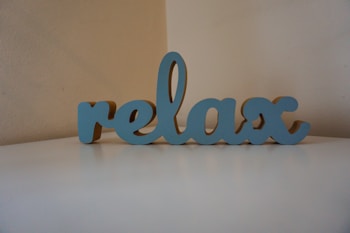 A blue wooden word sculpture spells 'relax' placed on a smooth white surface, against a simple beige wall background.