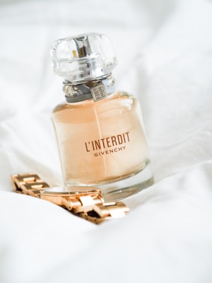 A bottle of Givenchy L'Interdit perfume with a metallic wristwatch placed next to it on a soft, white fabric background. The perfume bottle is elegant with a clear cap and contains a light amber liquid, while the watch has a shiny golden appearance.