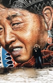 A large, detailed mural depicting the face of a person with expressive features painted on a brick wall. The mural includes vibrant colors, with emphasis on textures and shading. A person dressed in winter clothing, including a fur-trimmed hooded coat, stands smiling next to the mural, adding a sense of scale and interaction.