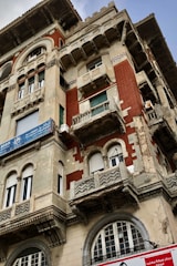 A detailed facade of an old building showcasing intricate architectural elements. The structure features ornate balconies, arched windows, and a combination of red brick and light stone materials. Signs with Arabic script are visible on the building, indicating commercial use. The wear on the facade suggests historical significance.
